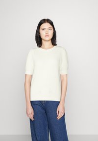 Šviesiai kreminis megztinis su trumpomis rankovėmis, raukšlėta apykakle ir apačia. Derinamas su laisvomis mėlynomis džinsomis. Lygus tekstūra, be raštų.