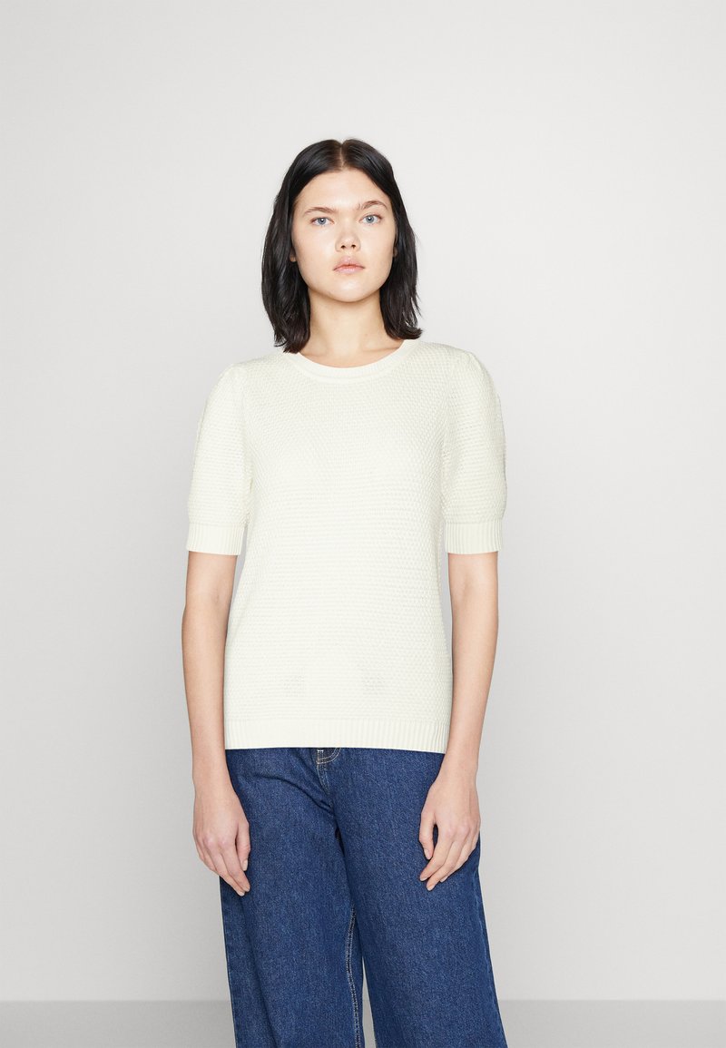 Šviesiai kreminis megztinis su trumpomis rankovėmis, raukšlėta apykakle ir apačia. Derinamas su laisvomis mėlynomis džinsomis. Lygus tekstūra, be raštų.