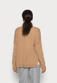 Suéter de punto beige con mangas largas, puños y dobladillo acanalados. Corte holgado con aberturas laterales, usado sobre una blusa de color claro y pantalones grises.