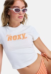 Witte kortemouwenshirt met oranje "ROXY 1990" logo, gecombineerd met gestreepte oranje en witte zwemkleding en witte cat-eye zonnebril.