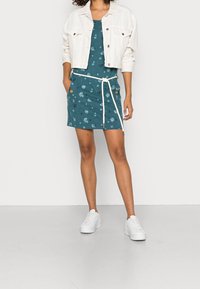 Aksamitna sukienka w kolorze turkusowym z kwiatowym wzorem, z regulowanym ściągaczem w talii i pomarańczowymi guzikami; połączona z białą dżinsową kurtką i białymi trampkami.