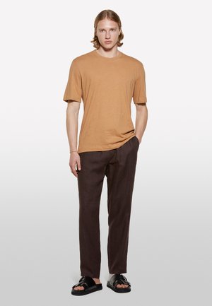 T-shirt a maniche corte di colore tan abbinato a pantaloni in lino marrone scuro e sandali neri. Il tessuto sembra morbido e leggermente testurizzato.