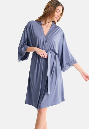 Woman tying belt on knee-length blue robe with lace trim on wide sleeves against plain light background.