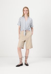 Pruhovaná košeľa s krátkym rukávom a uzlom vpredu, béžové strihané šortky a čierne lace-up baleríny. Jednoduchý, neformálny outfit na jednofarebnom pozadí.