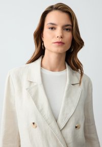 Hellbeige Leinen-Doppelreiher-Blazer mit Reverskragen, zwei Knöpfen und subtiler Textur. Getragen über einem weißen Rundhalsausschnitt-Shirt.