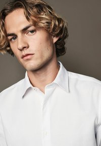 White textured button-up shirt with a pointed collar, featuring a single button at the neckline and a smooth finish.