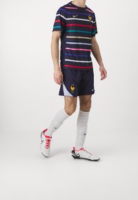 Mannen voetbaloutfit met een marineblauwe shirt met meerkleurige strepen en een hanenembleem, gecombineerd met marineblauwe shorts en witte sokken.