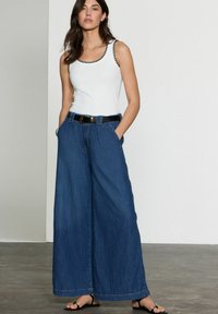 Vit linne med svart kant, i kombination med högmidjade, vida denimbyxor. Outfitsen kompletteras med ett svart bälte och sandaler.