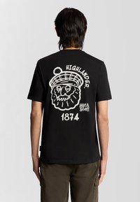 Sort bomulds t-shirt med et stort hvidt grafisk motiv på bagsiden, der viser en tegneseriefigur iført en tartan hat, tekst "HIGHLANDER" og "1874".
