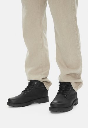 Czarne skórzane botki za kostkę z okrągłymi noskami, metalowymi oczkami i czarnymi sznurówkami. Podeszwy z teksturowanej gumy. Połączone z beżowymi, zwężanymi spodniami.