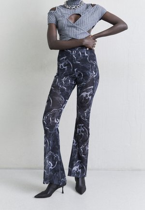 Šedý žebrovaný cropped top s výřezy na ramenou. Černé kalhoty s vysokým pasem, lesklým abstraktním vzorem a rozšířenými nohavicemi, doplněné o špičaté podpatky.