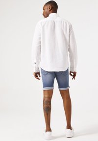 Chemise en lin blanc à manches longues, associée à un short en denim bleu retroussé. Coupe athlétique, texture subtile. Chaussures de sport blanches.