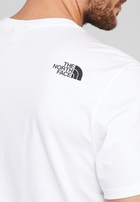 Hombre con una camiseta blanca con el logo negro de "The North Face" en la parte superior de la espalda, primer plano del área del hombro y el cuello.