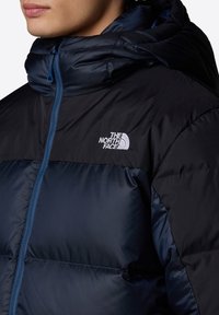 Une doudoune bleu marine foncé avec un col montant, une fermeture éclair latérale, et un logo brodé sur la poitrine, présentant une texture lisse et brillante.