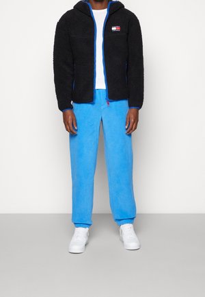 Zwarte fleecejack met blauwe accenten en capuchon, gecombineerd met felblauwe joggingsbroek van zacht materiaal, aangevuld met witte sneakers.