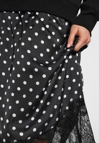 Jupe en satin noir à pois blancs, avec une bordure en dentelle à l'ourlet. Une main tient le tissu pour montrer sa texture et son mouvement.