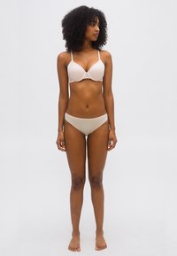 Γυναίκα με σγουρά μαλλιά, όρθια και ξυπόλητη, φορώντας μπεζ σουτιέν και εσώρουχο, κοιτάζει προς τα αριστερά της μπροστά σε απλό φωτεινό φόντο.