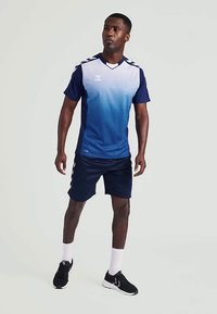 Mees seisab seljas sinise gradienti spordijersey, tumesinised lühikesed püksid, valged sokid ja mustad spordijalatsid, hallil taustal.