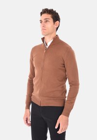 Maglione marrone con zip, realizzato in morbido tessuto a maglia, con colletto alto e polsini e bordo a costine, abbinato a pantaloni neri.