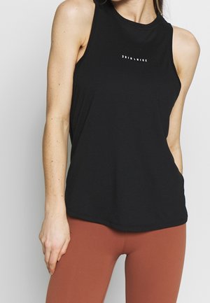 Frau trägt ein schwarzes ärmelloses Nike-Oberteil mit weißem Logo und rostfarbene Leggings, steht vor einem schlichten hellen Hintergrund.