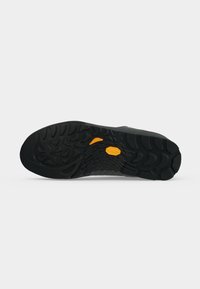 Semelle de chaussure noire avec des crampons profonds et deux petits patchs orange antidérapants conçus pour la traction et la durabilité.