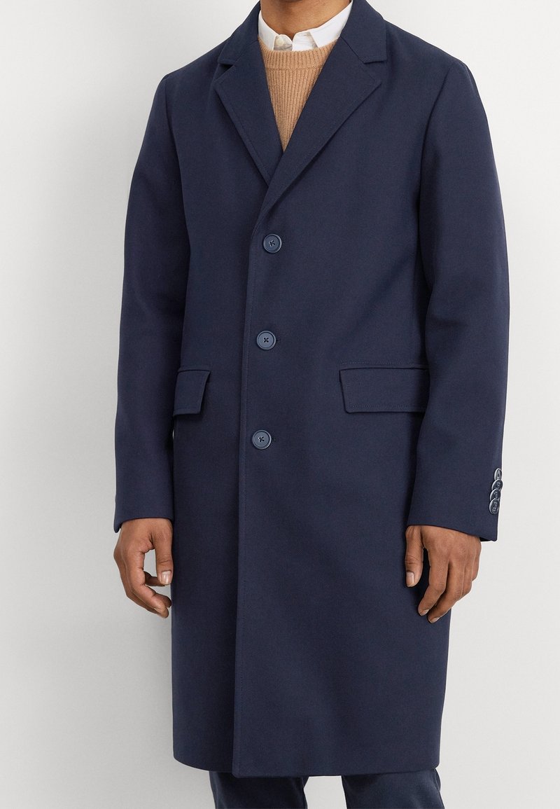 Marinblå ullkappa med rektangulär krage, tre framknappar, två sidofickor och knappade ärmslut. Slät textur och skräddarsydd passform.