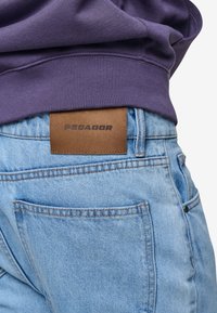 Světle modré džíny z denimu s viditelnými zadními kapsami a hnědou koženou nášivkou se slovem "PEGADOR" blízko pasu.