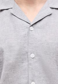 Ανοιχτό γκρι πουκάμισο με γιακά και τρία ορατά λευκά κουμπιά, φορεμένο από άτομο που εμφανίζεται από τον λαιμό μέχρι το μέσο του στήθους.