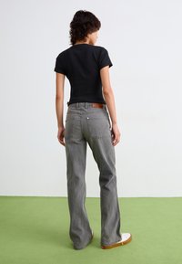 Camiseta negra de manga corta, jeans acampanados grises con bolsillos frontales y traseros, parche de cuero marrón en la cintura, zapatillas blancas, de pie sobre una superficie verde.