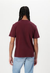Burgundy cotton t-shirt with a crew neck, short sleeves, and a straight hem. Features a small logo on the left sleeve.