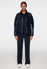 Granatowa puchowa kurtka z błyszczącym wykończeniem, poziomym pikowaniem, wysokim kołnierzem i przednim zamkiem błyskawicznym. Noszona na granatowych spodniach i białych butach.