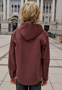 Veste à capuche bordeaux avec des coutures visibles, coupe décontractée et manches longues. Portée avec un jean foncé, vue de dos en extérieur.