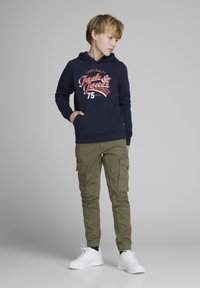 Laivasto huvtröja med vitt och rött logotyp, olivgrön cargobyxor och vita sneakers. Cargobyxorna har fickor och ribbade manschetter.