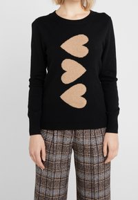 Schwarzer Pullover mit langen Ärmeln, der in der Mitte drei große beige Herzen aufweist, kombiniert mit braun-weißen karottenartigen Hosen.