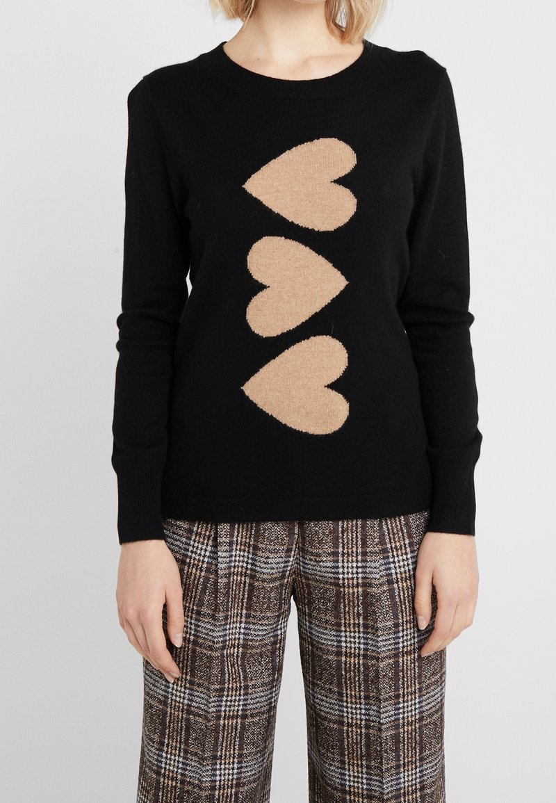Schwarzer Pullover mit langen Ärmeln, der in der Mitte drei große beige Herzen aufweist, kombiniert mit braun-weißen karottenartigen Hosen.