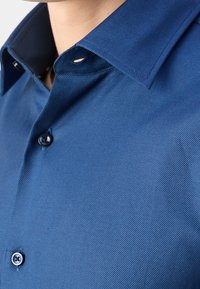 Niebieska koszula z teksturowanej tkaniny, kontrastowym ciemnym kołnierzykiem i zapięciem na guziki z widocznym jednym guzikiem na plisie.