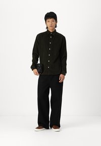 Mørkegrønn corduroy-skjorte med knappelukking, kombinert med svarte vide bukser og brune sneakers. Lite svart veske holdt i venstre hånd.