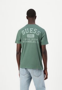 Zöld pamut póló, nagy fehér grafikával a hátán, rajta: "GUESS 1981 CLASSIC DESIGN LOS ANGELES, CA." Rövid ujjú, laza szabású.