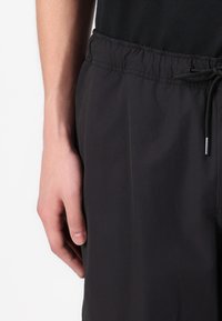 Black shorts with an elastic waistband and drawstring, featuring a smooth texture and a side pocket. Hand resting on the seam.