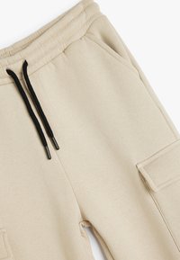 Beige Baumwoll-Jogginghosen mit schwarzen Kordeln, ausgestattet mit seitlichen Taschen und einer weichen Textur. Gerades Bein-Design mit minimalen Akzenten.