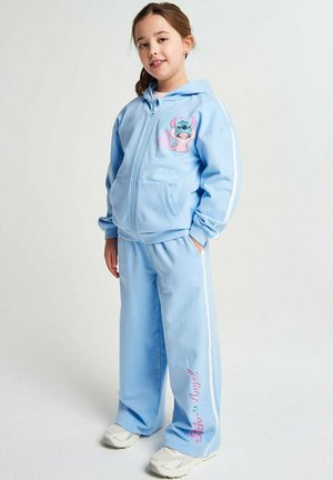 Young girl wearing light blue hooded tracksuit with cartoon character and "Sub & Angel" text, standing with hands in pockets.
