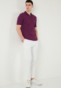 Lila galléros póló, rövid ujjakkal és félcipzárral, fehér, slim-fit nadrággal és fehér sportcipőkkel párosítva.