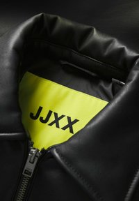 Kołnierz czarnej skórzanej kurtki z żółtym wnętrzem z napisem "JJXX" w czarnym kolorze. Posiada metalowy zamek błyskawiczny i gładką teksturę.