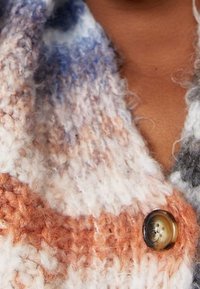 Pletený kardigan v měkké vícebarevné přízi s texturovaným povrchem. Obsahuje nadměrný hnědý knoflík a odstíny bílé, oranžové a modré.