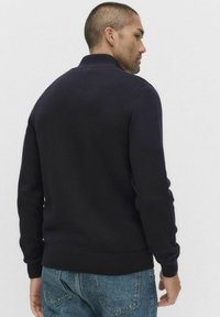 Granatowy sweter dziergany z wysokim kołnierzem, ściągaczami przy rękawach i u dołu. Wyróżnia się gładką fakturą, noszony z jasnoniebieskimi dżinsami.