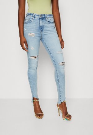 Femme portant un jean skinny déchiré bleu clair associé à des sandales à talons hauts colorées et un haut vert clair, debout devant un fond uni.