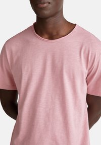 Hombre negro vistiendo una camiseta de cuello redondo de manga corta color rosa claro, de pie contra un fondo blanco liso con las manos detrás de la espalda.