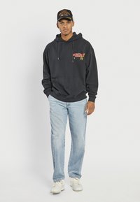 Schwarzer Kapuzenpullover mit grafischem Druck, kombiniert mit hellblauen Jeans. Modell trägt weiße Turnschuhe und eine schwarze Mütze mit roten Akzenten.