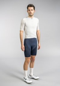 Men's cycling kit: fitted light gray jersey with short sleeves and a front zipper, paired with navy shorts featuring textured accents. White cycling shoes.