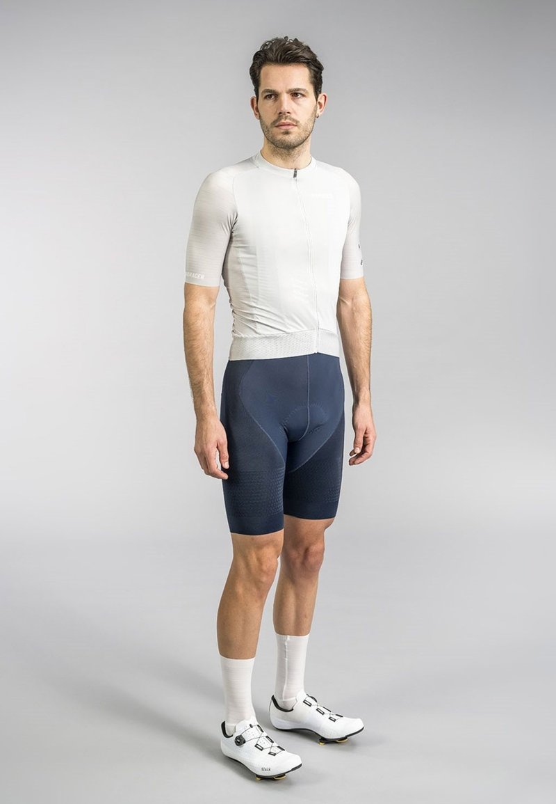 Men's cycling kit: fitted light gray jersey with short sleeves and a front zipper, paired with navy shorts featuring textured accents. White cycling shoes.